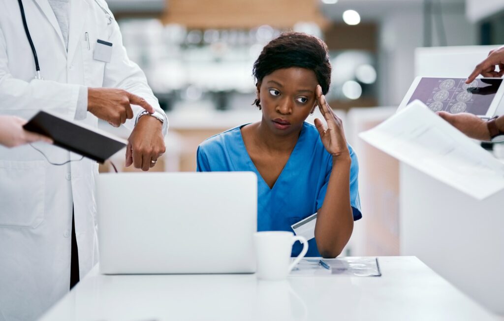 Stressed nurse sitting at her desk with laptop and coffee as multiple doctors approach her